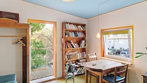 A room with a wooden bookshelf, a dining table with chairs, a window with a view of parked cars, and a hanging light fixture.