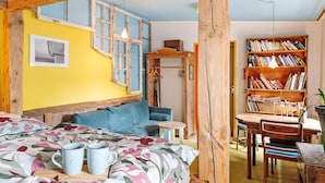 A room with a bed, a small table, a bookshelf, and a window with a view.