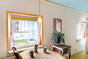 A room with a large window, a wooden table with a kettle and mugs, a potted plant, and a mirror on the wall.