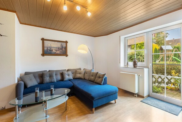 Ein Wohnzimmer mit Ecksofa, Glastisch, Stehlampe und einem großen Fenster mit Blick zum Garten.