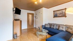 Un salon avec un canapé bleu, une table basse en verre, un téléviseur à écran plat, un meuble en bois et un tableau accroché au mur.