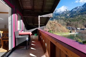 A balcony with a wooden railing, a wicker chair, a table, and a view of a lake and mountains.