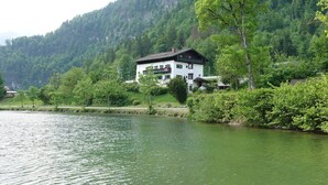 A two-story building with balconies, situated by a lake with a forested mountain backdrop.