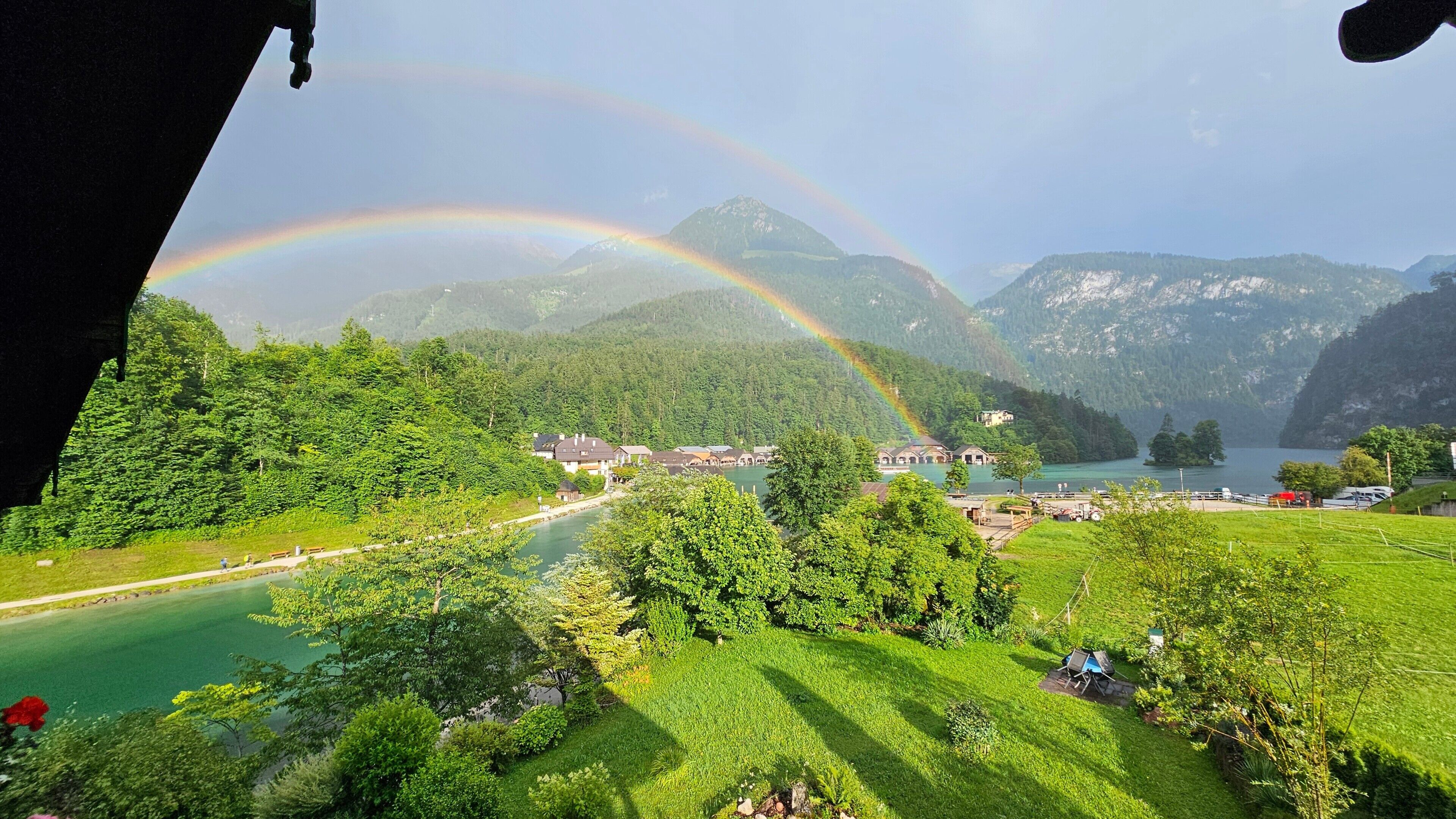 A scenic view of a lake surrounded by mountains, with a rainbow and a small village.