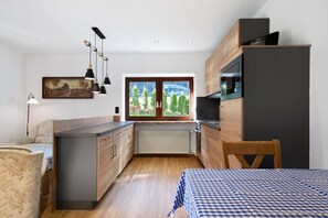 A compact kitchen with a view of trees and a building through a window, wooden cabinets, a dining area with a table and chairs, and a bed with a checkered tablecloth.