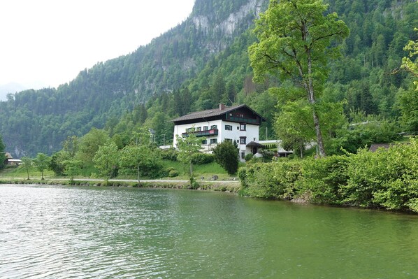 Un edificio de dos plantas con balcones, frente a un lago con montañas boscosas al fondo.