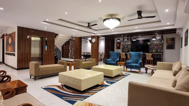 A modern living room with a sofa set, coffee table, and blue armchairs. There is a staircase with a wooden finish and a chandelier. - CALANGUTE BEACH VILLA By SEVEN HOTELS (Calangute)