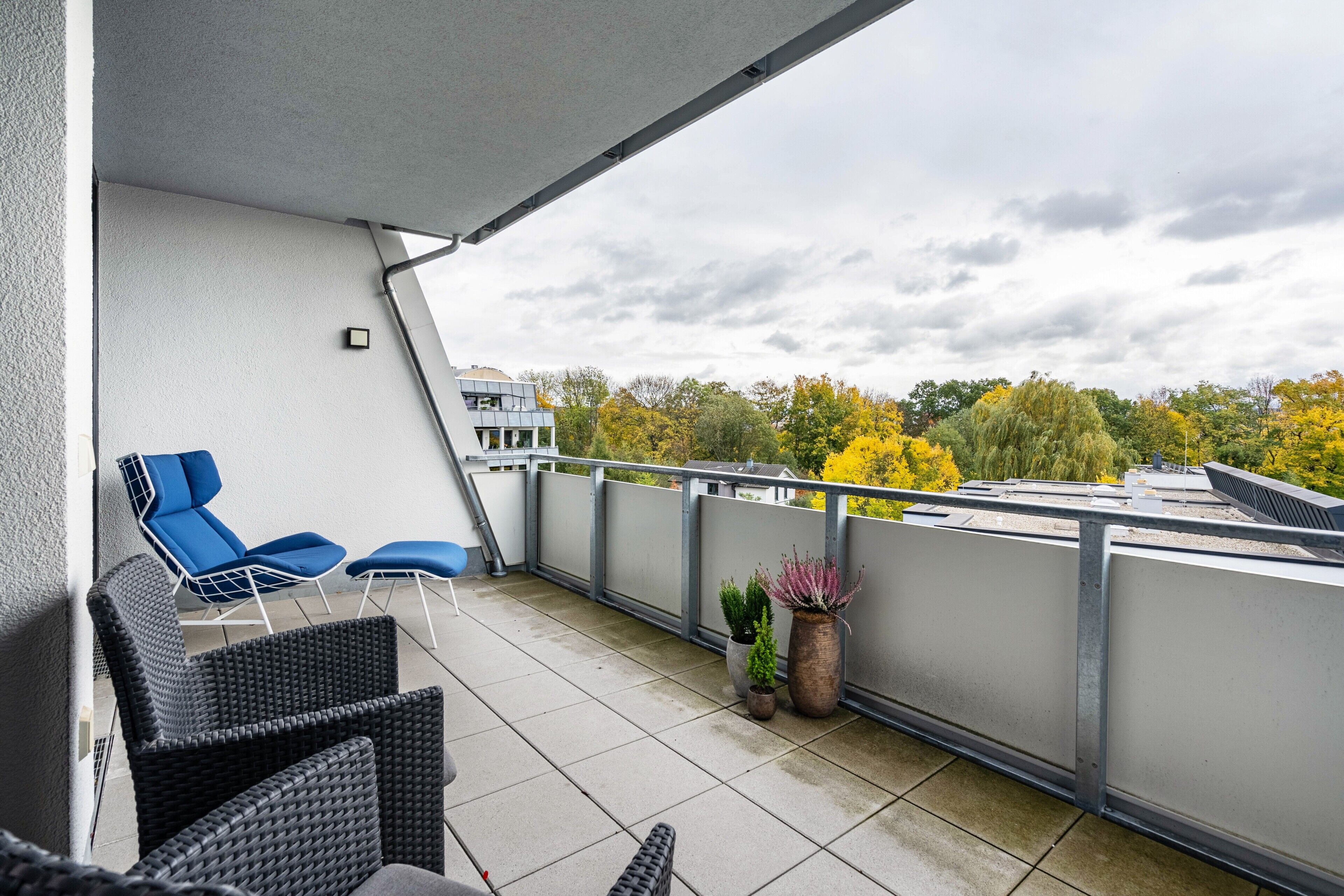 A balcony with outdoor seating, potted plants, and a view of trees and buildings.
