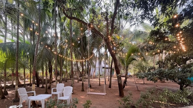 An outdoor seating area with white plastic chairs and tables, surrounded by trees and string lights. - Saaj Villa (Nagaon)