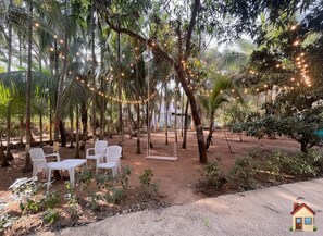 Un espace extérieur aménagé avec des chaises et des tables en plastique blanc, entouré d’arbres et illuminé de guirlandes lumineuses.
