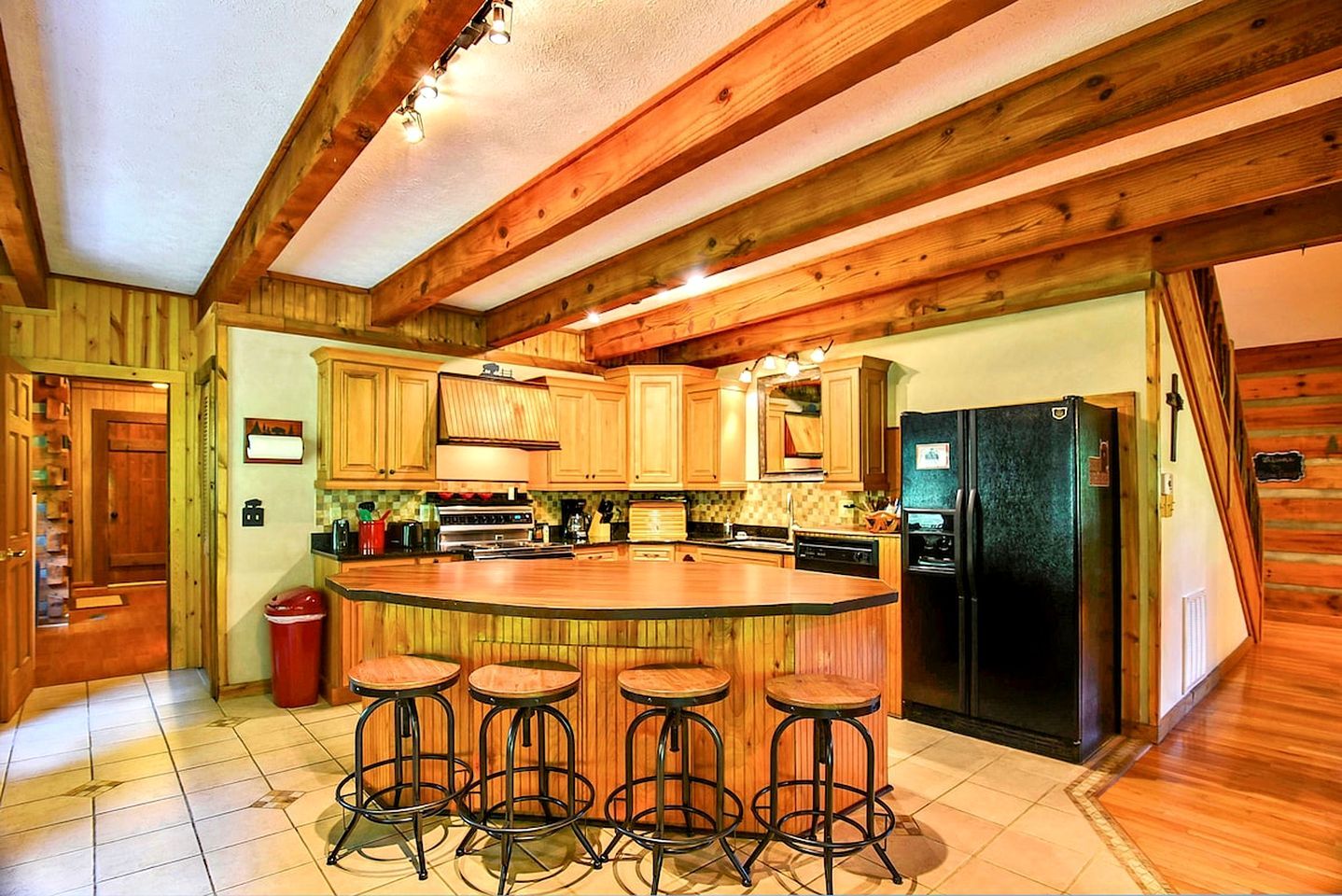 Una cocina con armarios de madera, una isla central con taburetes, un refrigerador negro y piso de baldosas.