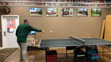 A person playing table tennis in a room with a ping pong table, a bookshelf with colorful items, and a sign indicating a nearby location.