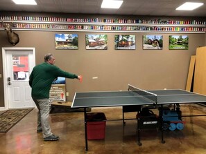 A person playing table tennis in a room with a ping pong table, a bookshelf with colorful items, and a sign indicating a nearby location. - Rustic Log Cabin Rental with a Hot Tub near Asheville, North Carolina (Clyde)