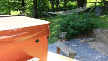 A hot tub with steps leading up to it, surrounded by a wooden deck and a grassy area with a hammock and a stone wall.