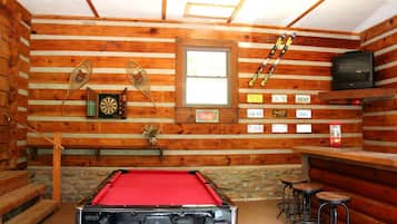 Interno di una baita in legno con tavolo da biliardo, freccette, televisore e targhe appese al muro.