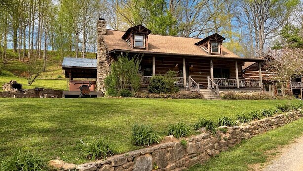 A log cabin with a stone chimney, a porch, and a stone retaining wall.
