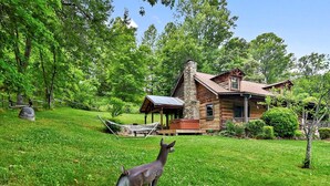 A rustic cabin with a stone chimney, surrounded by lush greenery and a hammock. - Rustic Log Cabin Rental with a Hot Tub near Asheville, North Carolina (Clyde)