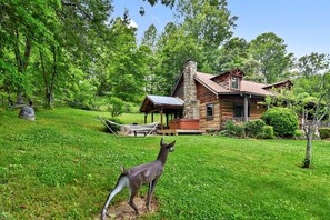 A rustic cabin with a stone chimney, surrounded by lush greenery and a hammock. - Rustic Log Cabin Rental with a Hot Tub near Asheville, North Carolina (Clyde)