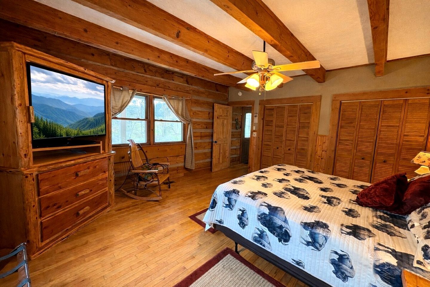 A log cabin bedroom with a bed, a television, a rocking chair, and a view of mountains through the window.