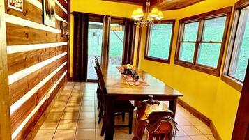 A wooden interior with a dining table and chairs, a chandelier, and large windows.
