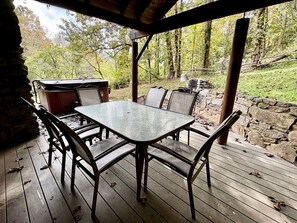 Une terrasse extérieure en bois, dotée d’une table à plateau de verre et de chaises en métal, entourée de murs en pierre et d’arbres.
