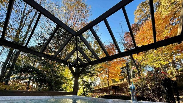 A hot tub under a wooden pergola with a view of autumn trees.