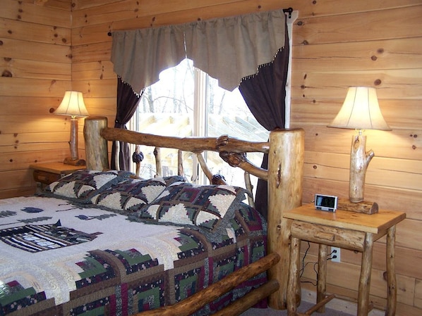 A wooden cabin bedroom with a bed, two bedside lamps, a clock, and a window with curtains. - Luxury Vacation Rental with a Hot Tub for Rent in Clyde, North Carolina (Clyde)