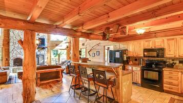 A wooden cabin interior with a stone fireplace, a kitchen with wooden cabinets, and a bar area with stools.