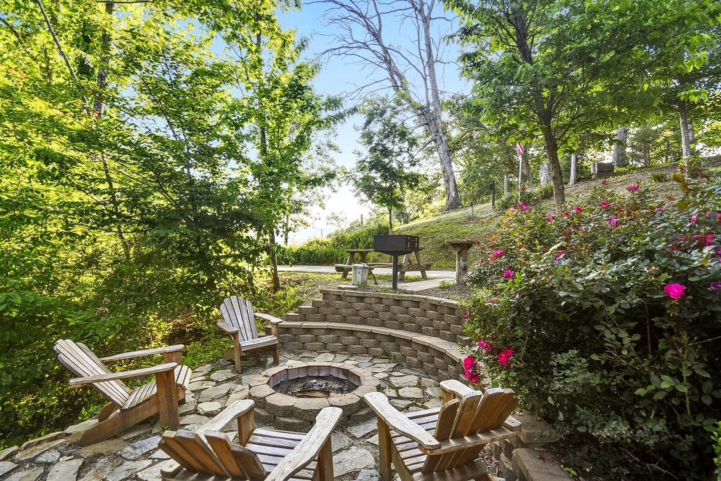A circular stone fire pit surrounded by wooden Adirondack chairs, set in a landscaped garden with trees and flowering bushes.