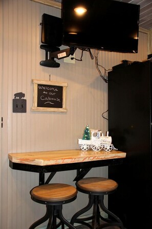 Un coin cuisine avec une table en bois, deux tabourets, un téléviseur fixé au-dessus de la table et une petite décoration de Noël.
