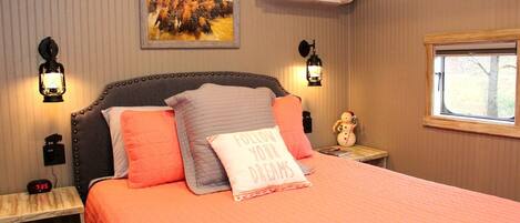 A bedroom with a bed, two bedside tables, a nightstand, a wall-mounted air conditioner, a window, and a painting on the wall.