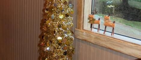 A room with a Christmas tree decorated with gold tinsel and ornaments, a wreath on the wall, and a window with a view of a tree and two small figures.