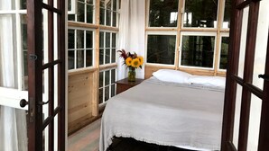 Een kamer met een bed, een houten bureau, een vaas met bloemen en grote ramen.