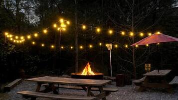 An outdoor setting with picnic tables, a fire pit, string lights, and an umbrella.