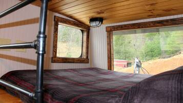 Un lit superposé avec une couverture à carreaux, une fenêtre donnant sur des arbres et un bâtiment, et un plafond en bois.
