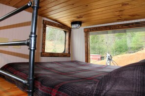 A bunk bed with a plaid blanket, a window with a view of trees and a building, and a wooden ceiling. - Glamping Caboose Rental Perfect for Families near Clyde, North Carolina (Clyde)