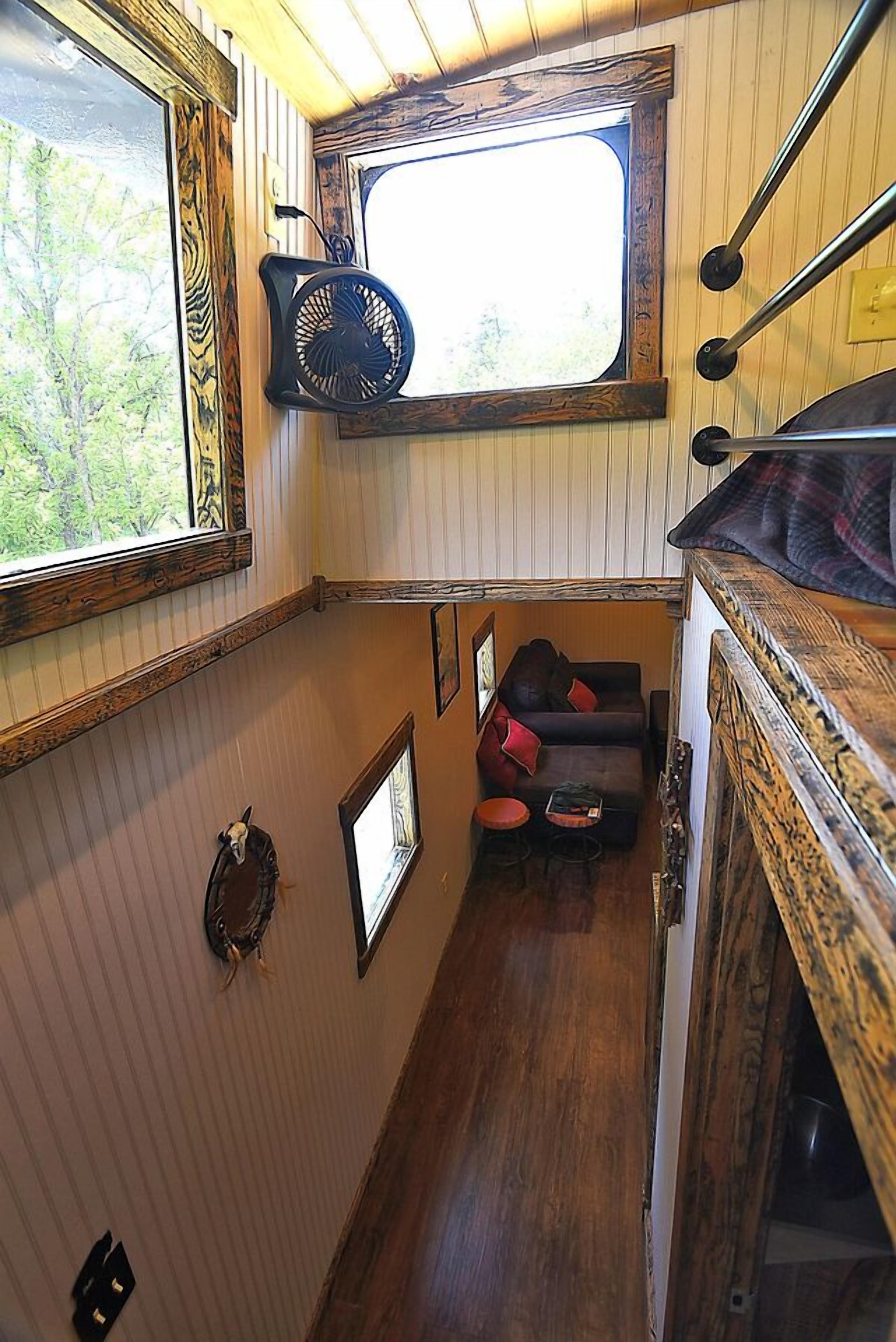 A narrow interior space with a wooden ceiling, a window with a view, a small seating area with a couch and pillows, and a fan mounted on the wall.