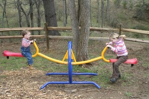 Two children playing on a seesaw in a wooded area with a wooden fence in the background. - Glamping Caboose Rental Perfect for Families near Clyde, North Carolina (Clyde)