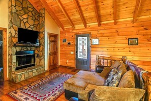 A log cabin interior with a stone fireplace, wooden walls, a television mounted above it, a door with a glass panel, and a sofa with patterned cushions.