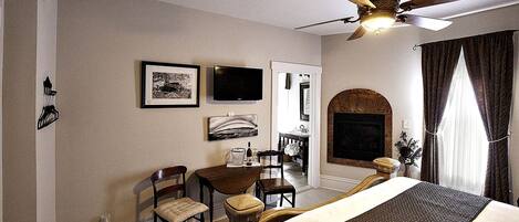 A hotel room with a bed, a desk, chairs, a television, a fireplace, and a ceiling fan.