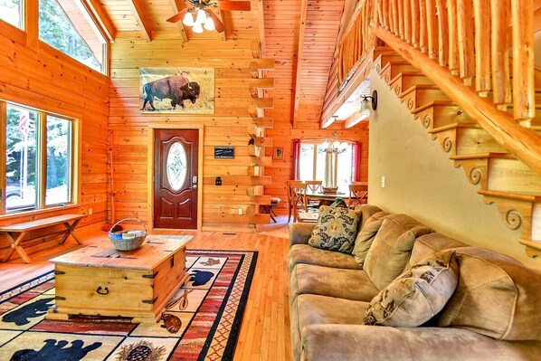 A log cabin interior with a wooden staircase, a dining area, and a living room with a sofa and a rug.