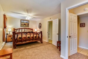 A bedroom with a wooden bed, a ceiling fan, a painting, and a door leading to another room.