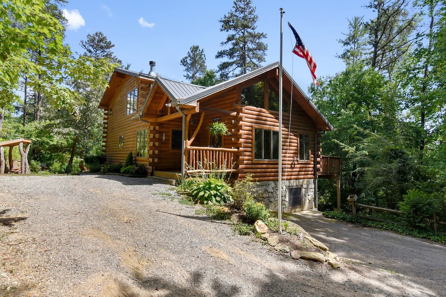 A log cabin with a porch, a flagpole with an American flag, and a gravel driveway.