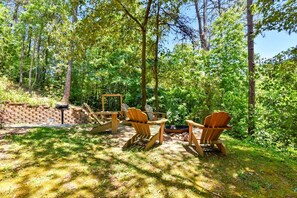 A forested outdoor area with Adirondack chairs and a fire pit.