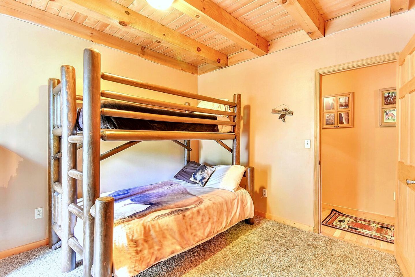 A bunk bed with a ladder, a single bed with a patterned blanket, a door with a mat, and a wall with framed pictures.