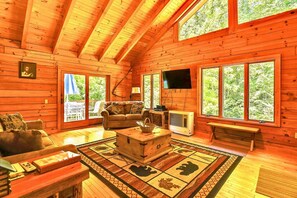A cozy cabin interior with a wooden floor, a sofa, a coffee table, and large windows offering a view of the forest.