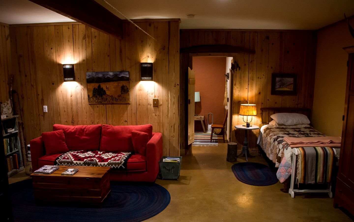 Una habitación acogedora con un sofá rojo, una mesita, una cama con colcha, paredes de madera y un cuadro en la pared.