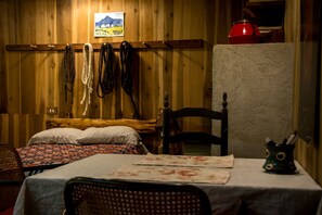 A wooden room with a bed, a chair, a table with a placemat, a red kettle, and a wall-mounted picture.