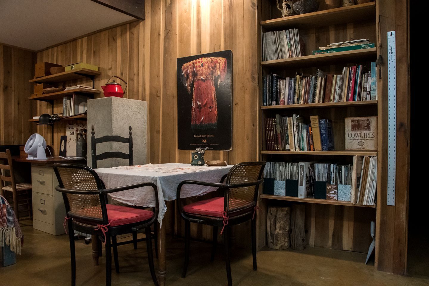 A room with a wooden wall, a bookshelf, a table with chairs, and a poster.