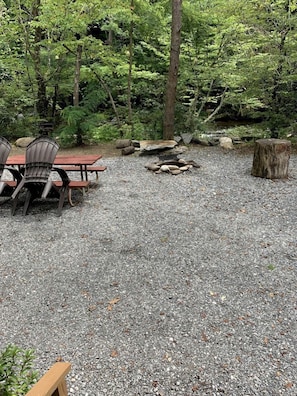 A gravel patio with a fire pit, wooden chairs, and a table, surrounded by trees.